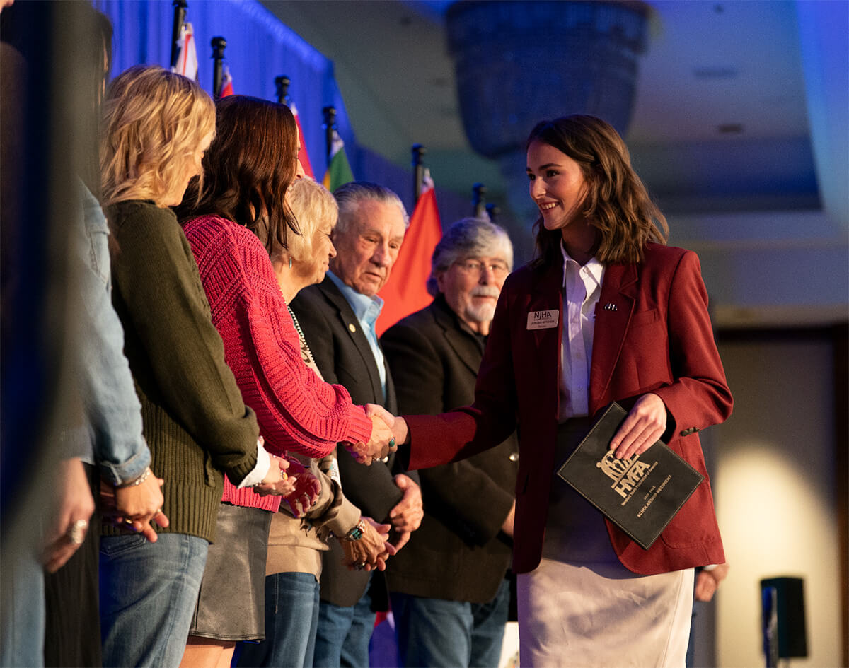 Woman in a red blazer holding an AHA folder shakes hands with an attendee onstage as a group stands beside flags.